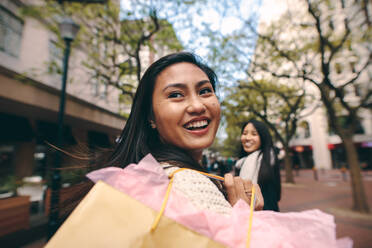 Two asian women walking in street carrying shopping bag. Rear view of a smiling woman carrying a shopping bag and looking back. - JLPSF23686