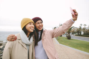 Zwei asiatische Frauen in Winterkleidung stehen am Meer und machen ein Selfie mit dem Handy. Freundinnen haben an einem kalten Tag Spaß im Freien. - JLPSF23692