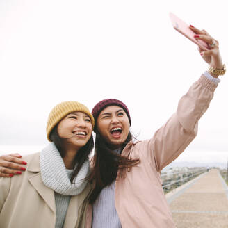 Lächelnde Freundinnen, die ein Selfie mit dem Handy im Freien machen. Fröhliche Freundinnen, die an einem kalten Tag in der Nähe des Meeres Spaß haben. - JLPSF23693