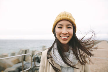 Nahaufnahme einer lächelnden Frau in Winterkleidung, die am Meer steht. Asiatische Frau, die an einem kalten Wintermorgen im Freien steht. - JLPSF23694