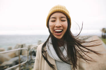 Fröhliche asiatische Frau, die in warmer Winterkleidung am Meer steht. Glückliche Frau, die im Freien steht und mit fliegenden Haaren Gesichter macht. - JLPSF23695