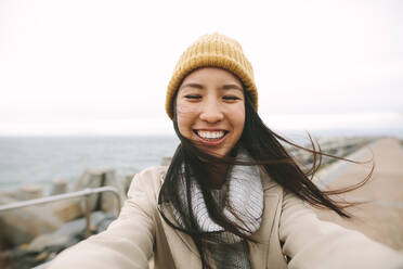 Close up of a smiling woman in winter wear standing near the sea. Asian woman standing outdoors on a cold winter morning with open arms. - JLPSF23696