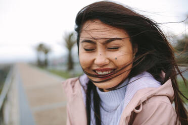 Close up of a woman standing outdoors with her hair flying on her face. Smiling woman standing on street enjoying the breeze with her eyes closed. - JLPSF23702