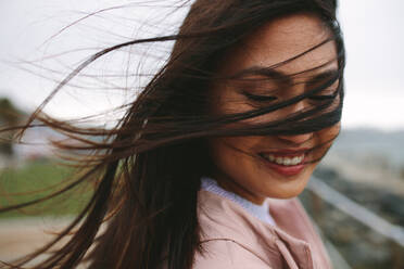 Lächelnde Frau, die auf der Straße steht und die Brise genießt, wobei ihre Haare fliegen. Porträt einer Frau, die im Freien steht und deren Haare ihr Gesicht verdecken. - JLPSF23707