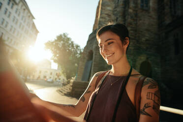 Lächelnde Frau, die an einer Straße steht und auf einen Stadtplan schaut, mit der Sonne im Hintergrund. - JLPSF23715