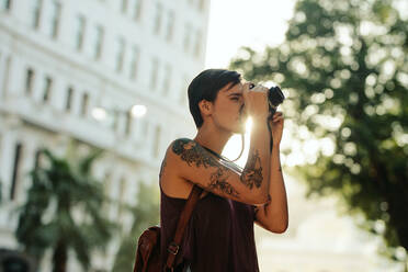 Frau Reisende fotografiert mit ihrer Kamera mit Sonne im Hintergrund. Tourist mit Tattoo auf der Hand, der durch die Stadt geht und fotografiert. - JLPSF23716