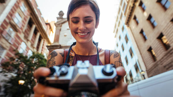 Nahaufnahme einer lächelnden Touristin, die mit einer Kamera fotografiert. Niedriger Winkel einer Frau, die ein Foto im Freien mit Gebäuden im Hintergrund macht. - JLPSF23725