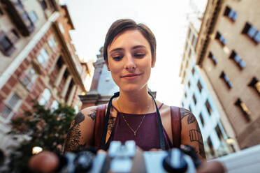 Nahaufnahme einer lächelnden Touristin, die mit einer Kamera fotografiert. Niedriger Winkel einer Frau, die ein Foto im Freien mit Gebäuden im Hintergrund macht. - JLPSF23726