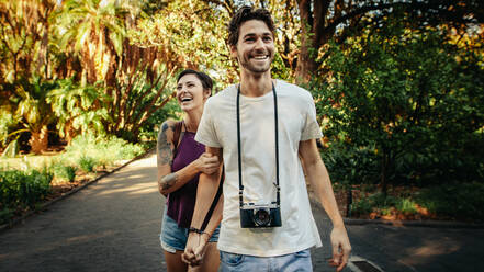 Glückliches Entdeckerpaar, das auf einem Weg in einem Park spazieren geht. Der Mann trägt eine Kamera und die Frau hält ihre Hand. - JLPSF23730