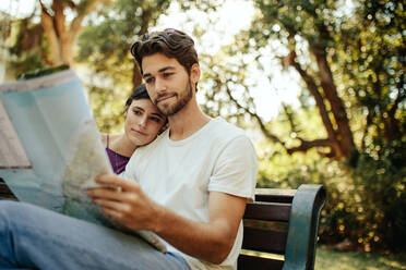 Tourist couple using a map to find the route to their destination. Man and woman travelers sitting on a bench in a park looking at a map. - JLPSF23734