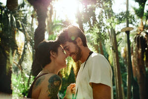 Mann und Frau stehen eng beieinander und berühren ihre Nasen in einem Park mit Sonne im Hintergrund. Touristenpaar in einer glücklichen und romantischen Stimmung bei der Erkundung der Stadt. - JLPSF23735