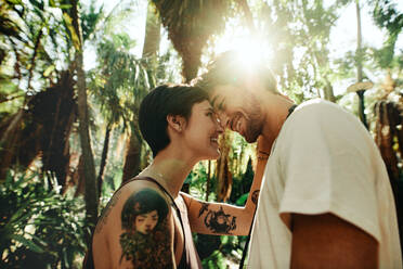 Mann und Frau stehen dicht beieinander und berühren ihre Stirne in einem Park mit hohen Bäumen und Sonne im Hintergrund. Touristenpaar in einer glücklichen und romantischen Stimmung, während sie die Stadt erkunden. - JLPSF23737