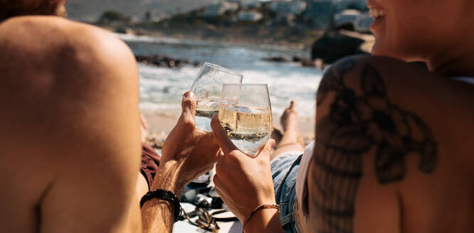 Nahaufnahme von hinten eines Touristenpaares, das am Strand sitzend Getränke in Gläsern hält. Paar im Urlaub, das am Strand sitzend auf seine Getränke anstößt. - JLPSF23744