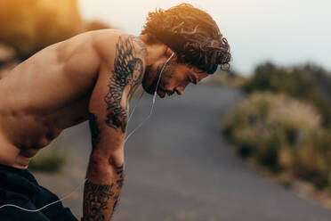 Seitenansicht eines muskulösen Mannes, der sich nach einem Lauf im Freien ausruht. Sportler, der nach einem Lauftraining auf einer Bergstraße müde aussieht. - JLPSF23747