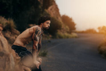 Sportsman relaxing after his morning workout in open air. Athlete looking tired and exhausted after morning run on mountain road. Man leaning to a rock by the road. - JLPSF23748