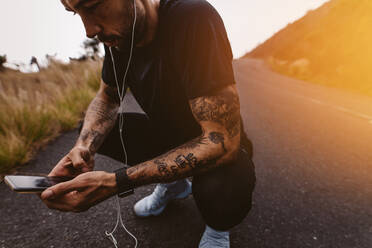 Close up of runner crouching on a mountain road and looking at his mobile phone. Athlete with earphones checking is fitness progress on smartphone app during workout break. - JLPSF23749