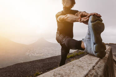 Male athlete stretching his leg after morning run on mountain road. Sports man stretching muscles outdoors after running. Focus on shoe. - JLPSF23750