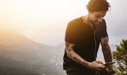 Young man using mobile phone while having exercise break outdoors in nature. Fit sportsman with smartphone checking his fitness progress. - JLPSF23751