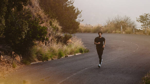 Horizontalaufnahme eines Läufers, der für einen Marathonlauf trainiert. Ein Mann läuft morgens auf einer Bergstraße bergab. - JLPSF23764