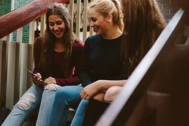 Drei junge Frauen sitzen auf der Treppe und schauen auf ihr Handy. Eine Gruppe von Highschool-Mädchen sitzt auf der Treppe. - JLPSF23768