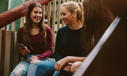 Junge Frauen sitzen in der Pause auf einer Treppe in der Universität. Drei Highschool-Mädchen sitzen auf einer Treppe im Freien mit einem Mobiltelefon. - JLPSF23769