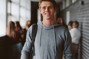Porträt eines glücklichen jungen Mannes mit Tasche, der in einem Universitätskorridor mit Klassenkameraden im Hintergrund spazieren geht. Junger männlicher Student auf dem College-Campus. - JLPSF23775