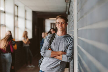 Porträt eines gut aussehenden jungen Universitätsstudenten, der sich an eine Wand in einem College-Korridor lehnt, mit anderen Studenten im Hintergrund. Kaukasischer männlicher Student auf einem High-School-Campus. - JLPSF23776