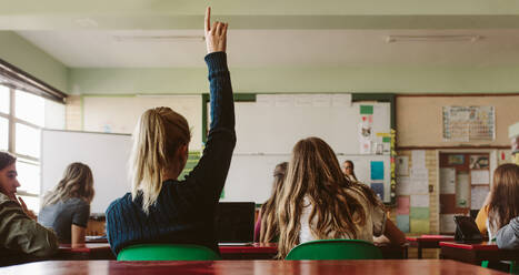 Rear view of female student sitting in the class and raising hand up to ask question during lecture. High school student raises hand and asks lecturer a question. - JLPSF23785