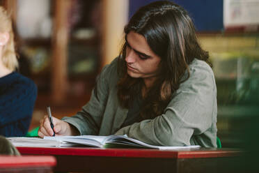 Junger Mann schreibt während einer Vorlesung Notizen in ein Buch. Männlicher Student studiert im Klassenzimmer. - JLPSF23789
