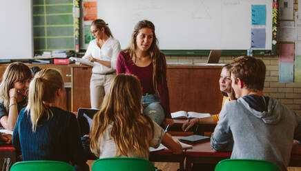 Junge Frau sitzt am Klassentisch mit Klassenkameraden um sich herum und unterhält sich. Gruppe von Gymnasiasten bespricht neue Aufgaben im Klassenzimmer. - JLPSF23804