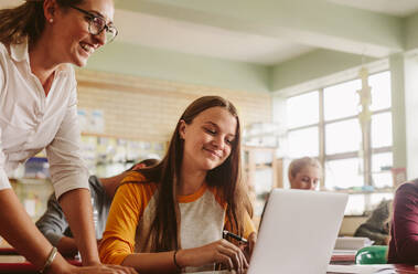 Junge Frau, die an einem Laptop lernt, während ein Lehrer im Klassenzimmer steht. Ein Highschool-Lehrer hilft einem Schüler im Klassenzimmer. - JLPSF23807