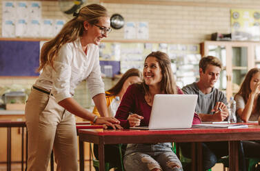 Glückliche Frau, die als Highschool-Professorin den Schülern während ihres Unterrichts hilft. Gruppe junger Leute im Klassenzimmer mit hilfsbereiter Lehrerin. - JLPSF23808