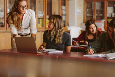 Teacher talking to female student sitting at the desk with book and laptop. Teacher with students during her class in high school classroom. - JLPSF23815