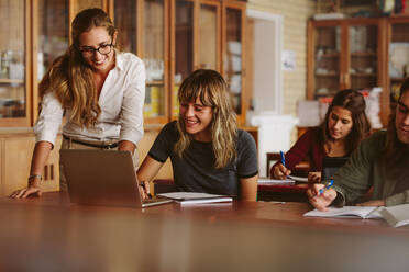 Glückliche Lehrerin, die ihrem Schüler in der High School hilft. Frau, die einen Laptop benutzt, während ihr Lehrer im Klassenzimmer steht und lächelt. - JLPSF23816