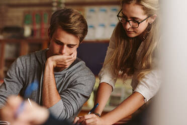 College-Student sitzt an seinem Schreibtisch mit einer Professorin, die sein Notebook überprüft. Vorlesung, die einem Studenten während ihrer Vorlesung hilft. - JLPSF23822