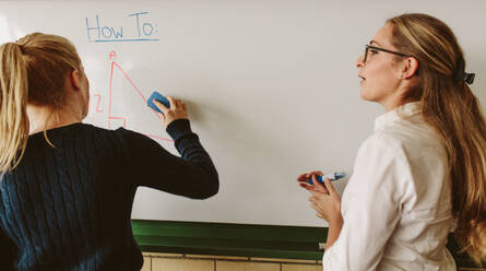 Rear view of young student writing on board with teacher standing by in classroom. Student writing on board during geometry class. - JLPSF23829