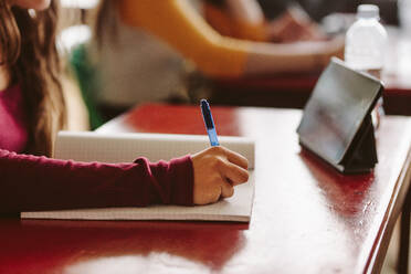 Nahaufnahme einer Studentin, die mit der Hand in ein Notizbuch mit digitalem Tablet auf dem Schreibtisch schreibt. - JLPSF23836