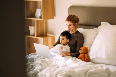 Young mother with her beloved son in the bedroom on the bed watching a movie on laptop. Adorable boy sitting on bed with mother and watching cartoons on laptop computer. - JLPSF23845