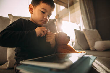 Kleiner Junge sitzt mit digitalem Tablet auf dem Sofa und isst Minzbonbons. Unschuldiges Kind zu Hause mit digitalem Tablet. - JLPSF23849