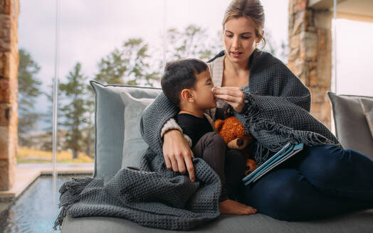 Mutter putzt ihrem kleinen Sohn die Nase. Mutter und Sohn sitzen auf dem Sofa, eingewickelt in eine Decke und mit einem digitalen Tablet. Die Mutter hilft ihrem Sohn, sich die Nase zu putzen. - JLPSF23850