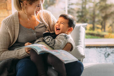 Mutter und Sohn schauen sich an, halten ein Buch in der Hand und lachen, während sie zu Hause auf der Couch sitzen. Glückliche junge Familie, die Spaß beim Lernen hat. - JLPSF23856