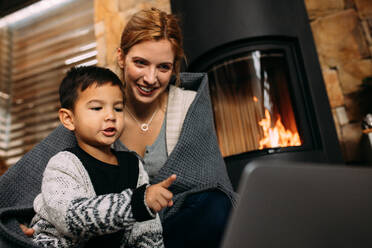 Little boy pointing at laptop while sitting with mother near fireplace at home. Mother and son looking at something interesting on laptop computer in living room. - JLPSF23858