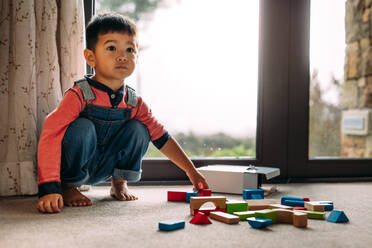 Unschuldiges kleines Kind sitzt auf dem Boden und hat bunte Holzklötze vor sich liegen. Kleiner Junge spielt mit Spielzeug zu Hause. - JLPSF23866