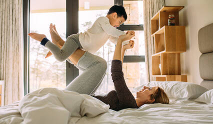 Seitenansicht einer glücklichen jungen Frau, die mit einem kleinen Kind auf dem Bett spielt. Eine Frau liegt auf dem Bett und hebt ihren Sohn mit ihren Beinen hoch. - JLPSF23875