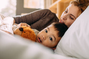 Süßer kleiner Junge hält seinen Teddybär und liegt mit seiner Mutter auf dem Bett. Kleines Kind im Bett mit seinem Spielzeug und seiner Mutter. - JLPSF23878