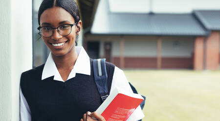 Teenager-Mädchen, das ein Lehrbuch in der Hand hält und durch den Schulkorridor geht. Selbstbewusste Schülerin in Uniform, die in die Kamera schaut und lächelt, auf dem Weg zum Unterricht. - JLPSF23881
