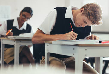 Teenage boy is taking a test while sitting at desk in classroom in high school. Students are concentrating while writing during exam. - JLPSF23887