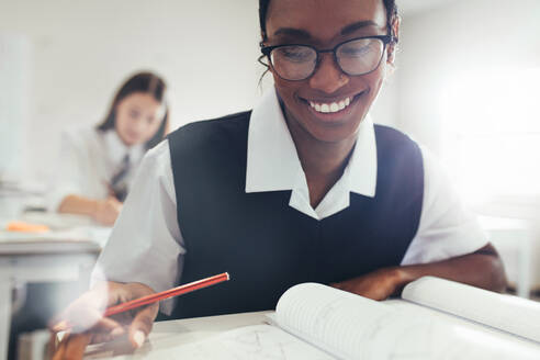 Teenager-Mädchen mit Brille sitzt am Schreibtisch im Klassenzimmer in der High School. Studentin lächelnd beim Lernen im Klassenzimmer. - JLPSF23888