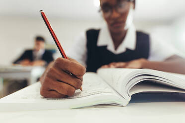 Nahaufnahme einer Teenagerin, die in ihr Lehrbuch schreibt, während sie am Schreibtisch im Klassenzimmer sitzt. High-School-Schülerin konzentriert sich beim Schreiben in einem Buch, Fokus auf die Hand der Schülerin. - JLPSF23889