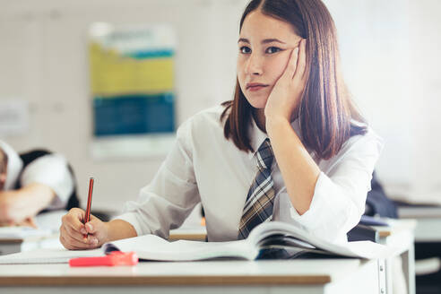 Teenager-Mädchen in Uniform sitzt am Schreibtisch im Klassenzimmer und denkt. Weibliche Studentin denkt beim Lernen im Klassenzimmer. - JLPSF23894